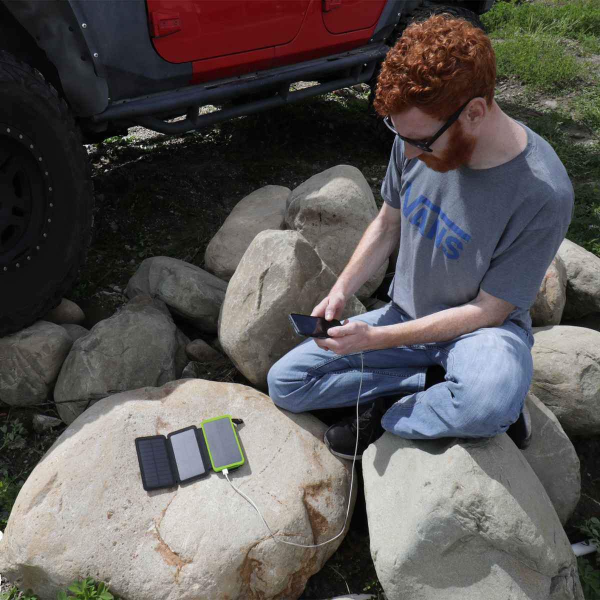 A person charging their smartphone with the JackPak PB20KS solar power bank while sitting on rocks outdoors next to a red vehicle.