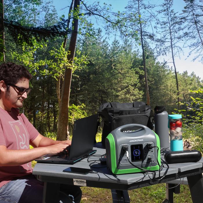 Man working on a laptop outdoors with JackPak PS600W power station charging devices, including a phone and laptop, in a forest setting.