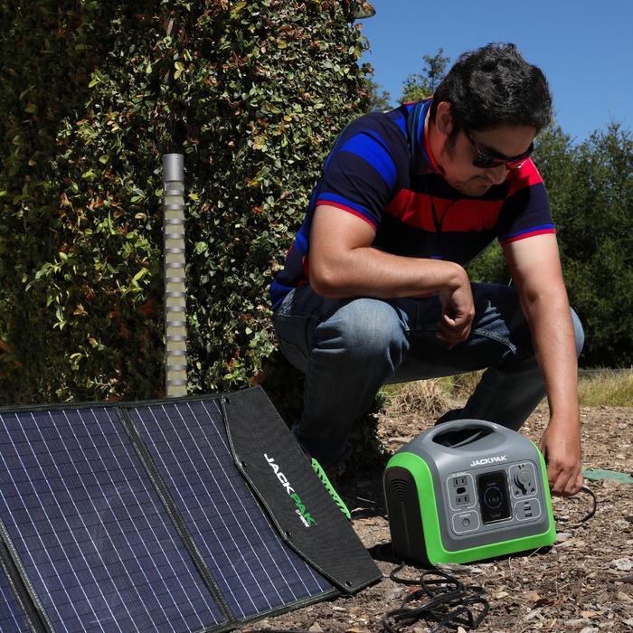 A dapper young gentleman crouching outdoors, connecting the JackPak PS600W portable power station to a JackPak solar panel for charging. The power station is positioned on the ground, with the solar panel next to it, showing the versatility of renewable e